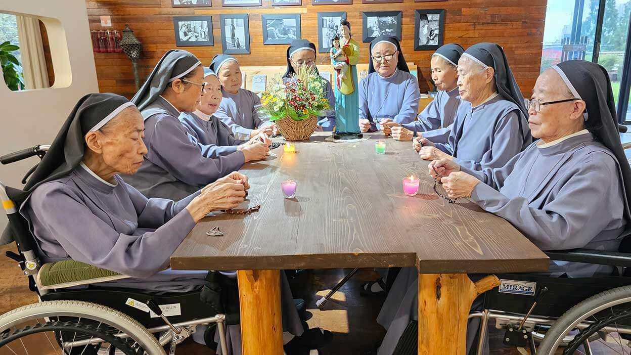 Elderly Sisters of Mary Sisters seated around a table, praying together in community.
