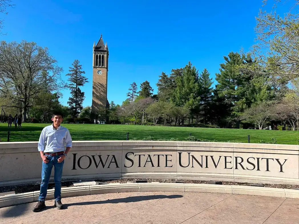 Sisters of Mary graduate Orlin stands near the Iowa State University sign with the campus bell tower behind him.